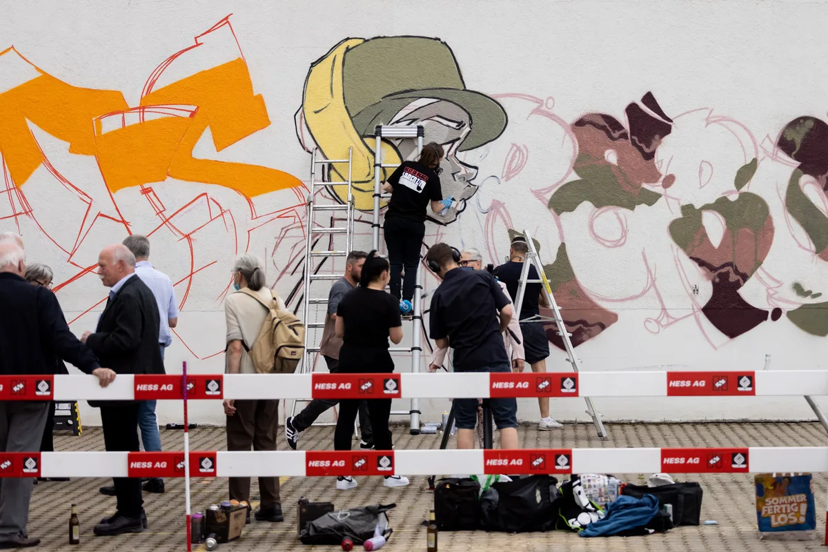 Man sieht Menschen hinter einer Bauabschrankungen. Im Hintergrund ist eine Sprayerin dabei, ein Kunstwerk an die Wand zu sprühen.